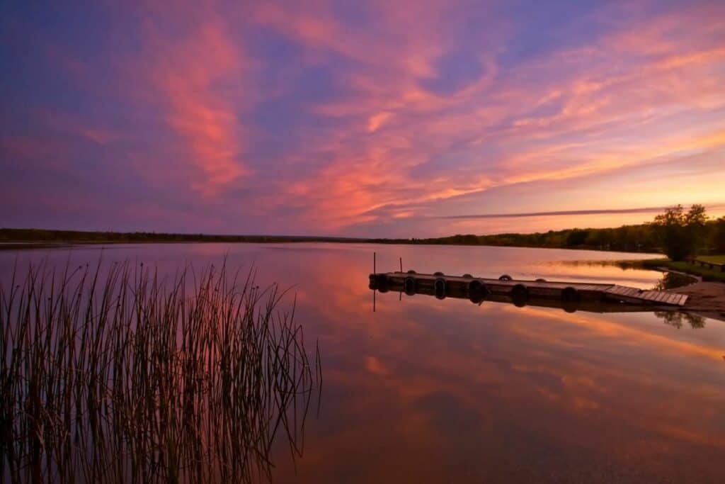 A Sunrise Of Orange, Red And Purple Over A Manitoba Lake With Reeds In The Foreground Is One Of The Best Reasons To Move To Manitoba